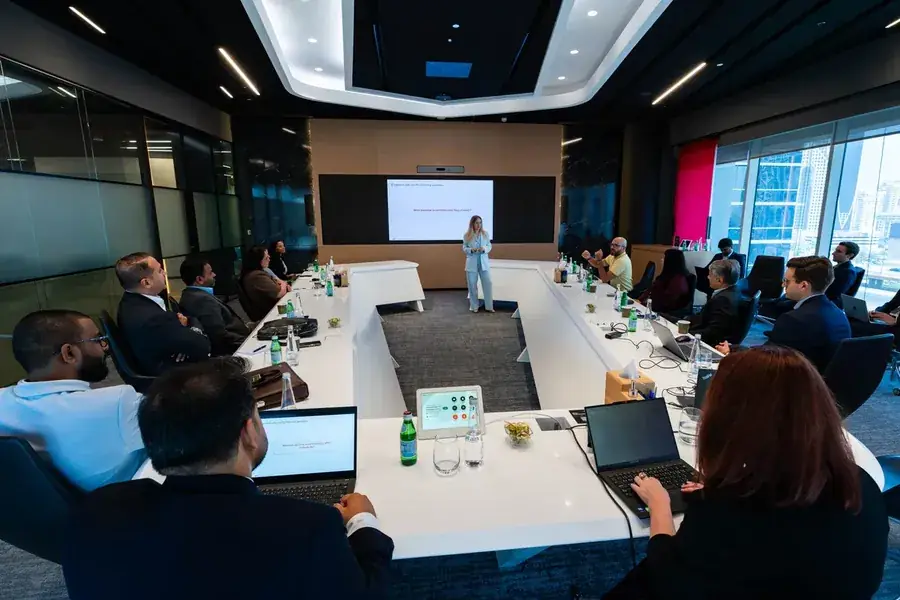 Kate V. presenting to a boardroom of seated attendees around a large table, a slide on the screen behind her.