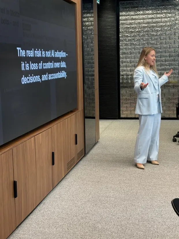 Kate V. in a pale blue suit presenting beside a large screen that reads: the real risk is not AI adoption, it is loss of control over data, decisions, and accountability.